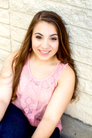 Authentic senior portrait of young woman in a pink shirt sitting next to a white stone wall.